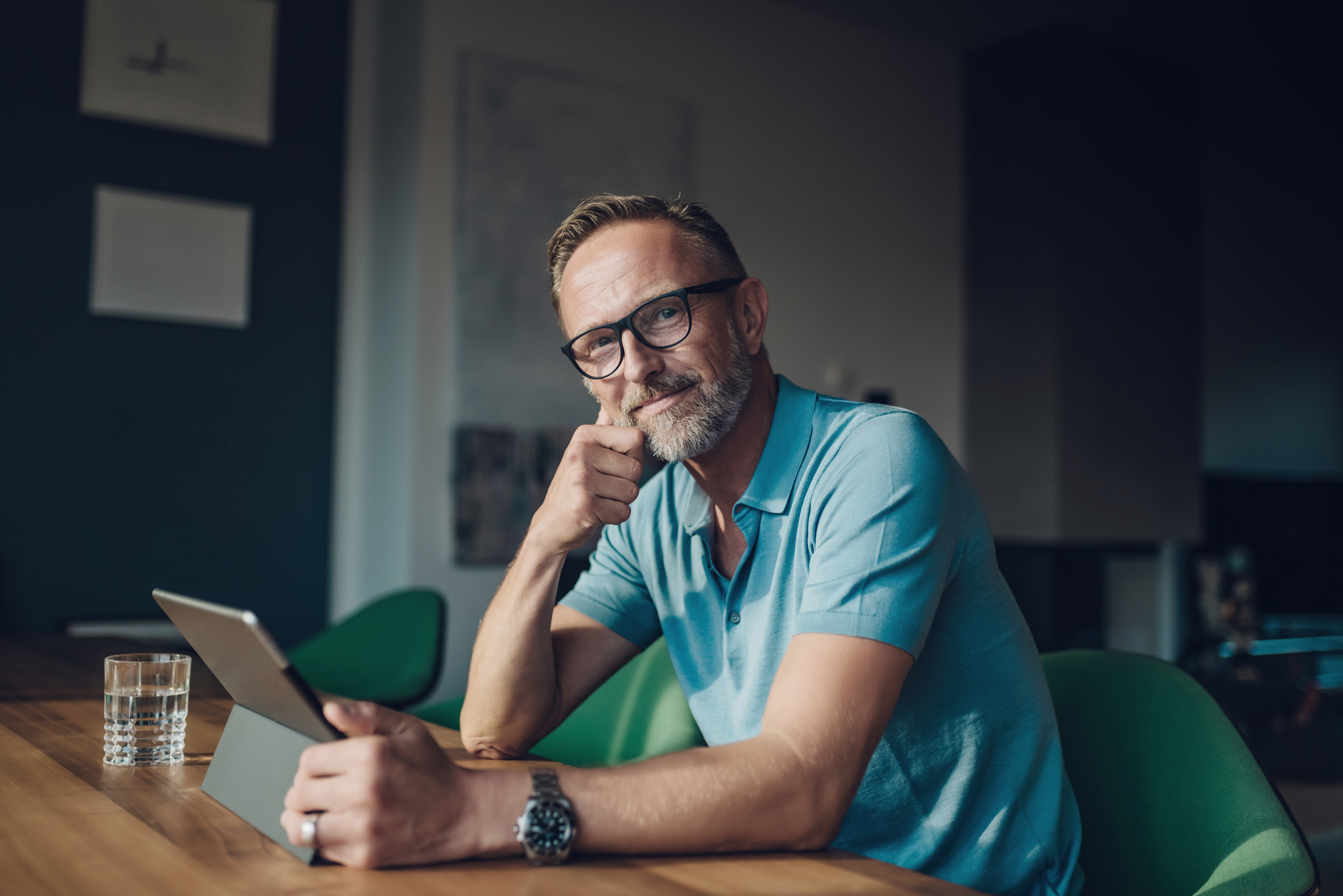 A man is sitting at a table indoors, using a tablet. He is wearing clothing that suggests casual or professional attire. The setting includes a wall in the background and furniture around him, with additional details like a watch and glasses visible.